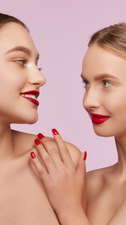 Portrait of two young, beautiful girls with well-kept skin and red lips makeup isolated over pink studio background. Concept of skincare, cosmetology, natural beautyの写真素材