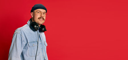 Portrait of man in stylish modern clothes, glasses, hat and headphones posing over red background....