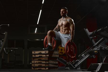 Portrait of young muscular man training with weights, posing shirtless in gym indoors. Relief body...