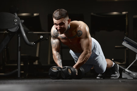Endurance. Portrait of young muscular man training shirtless in gym indoors. Doing push ups with dumbbells. Concept of sport, workout, strengthの写真素材