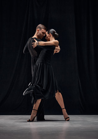 Sensual dance type. Man and woman, professional tango dancers performing in black stage costumes over black background.の写真素材