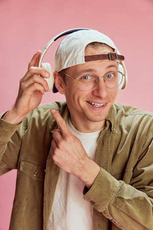 Good song. Young positive man in cap and glasses posing, listening to music in headphones over pink...