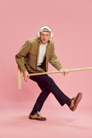 Young emotional man posing, listening to music in headphones, playing with mop like guitar over pink...