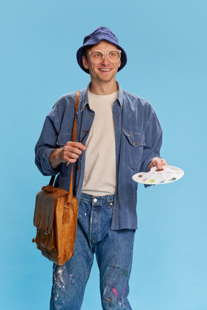 Positive mood, inspired look. Portrait of young man in jeans clothes, painter, artist posing over blue studio background. Concept of emotions, hobby, occupationの写真素材