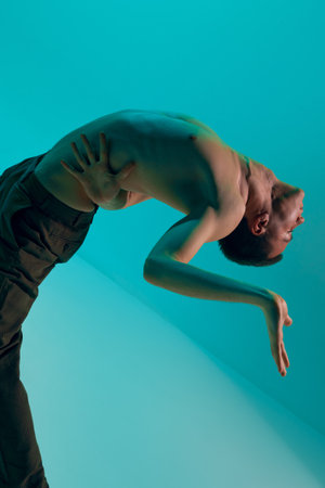 Contemporary dance style. Young flexible shirtless man dancing experimental dance over cyan color...