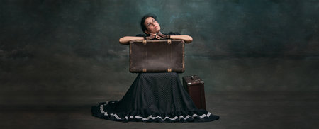 Arriving. Beautiful woman in image of Anna Karenina sitting on retro suitcase over vintage dark...