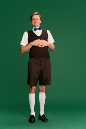 Positive. Portrait of man in stylish classical clothes, shorts and vest posing over green studio background. Concept of emotions, facial expression, lifestyleの写真素材