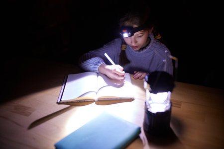 Little school girl sitting at table at home in the evening and doing homework without electricity...