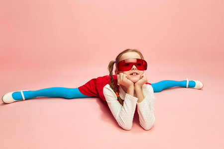 Girl, child in red dress, blue tights and red glasses sitting on twine, posing over pink studio background. Concept of emotions, childhoodの写真素材