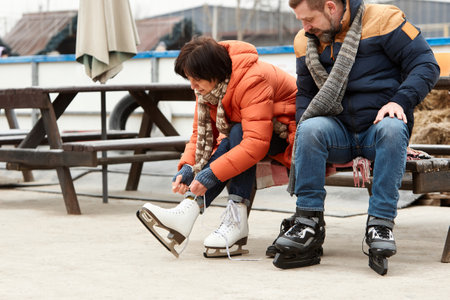 Mature man and woman getting ready to skate on ice rink outdoors. Woman tying laces on white skates. Active weekendsの写真素材