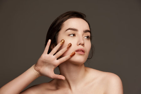 Sparkling nude eyeshadow swatches on fingers. Portrait of young beautiful girl with brown hair posing over dark grey studio background. Concept of natural beauty, skin care, cosmetologyの写真素材