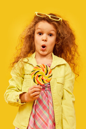 Shocked face. Little cute girl, child with curly hair posing in bright clothes with candy over yellow studio background. Concept of childhood, emotions, funの写真素材