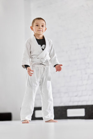 Portrait of little boy, child in white kimono attending sport class, training judo, jiu-jitsu in...