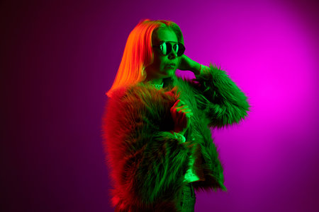 Trendy look. Stylish young woman posing in fur coat and sunglasses over magenta studio background in green neon light. Concept of emotions, lifestyle, fashionの写真素材