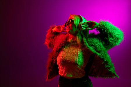 Young stylish woman posing in fur coat, listening to music in headphones over magenta studio background in green neon light. Concept of emotions, lifestyleの写真素材