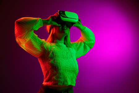 Modern gadgets and technologies. Young woman in VR glasses over magenta studio background in green...