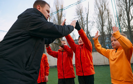 Professional football coach giving high five, cheering kids football team on field outdoor....