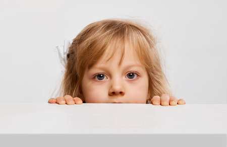 Curious kid. Cute little girl, child peeking out table, looking against grey studio background. Concept of childhoodの写真素材