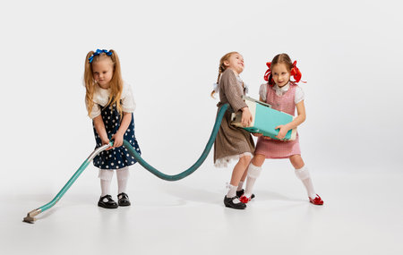 Little beautiful girls, kids in retro dresses playing with vacuum cleaner against grey studio background. Concept of childhoodの写真素材