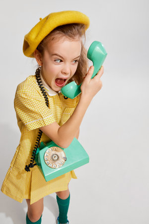Beautiful little girl, kid in retro yellow dress emotionally talking on phone, posing against grey studio background. Concept of childhoodの写真素材