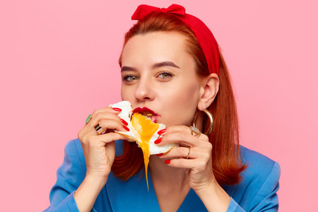 Close-up portrait of young redhead woman eating fried eggs against pink studio background. Liquid yolk. Food pop art photography.の写真素材
