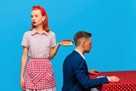 Young redhead woman serving breakfast with fried eggs and sausages to man against blue studio background. Food pop art photography.の写真素材