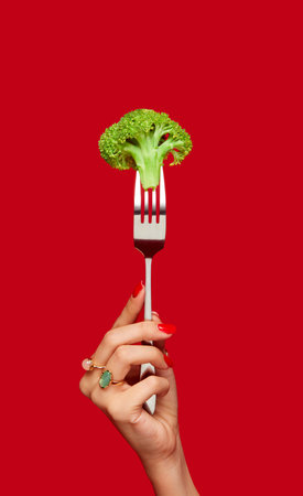 Female hand holding broccoli on fork against red studio background. Healthy eating. Food pop art photography.の写真素材