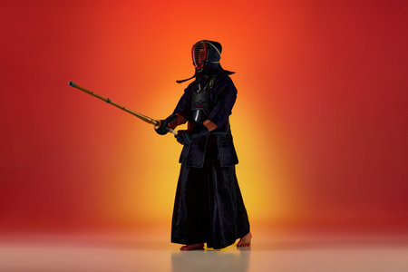 Full-length portrait of man, professional kendo athlete in uniform posing with shinai sword against gradient studio background in neon lightの写真素材