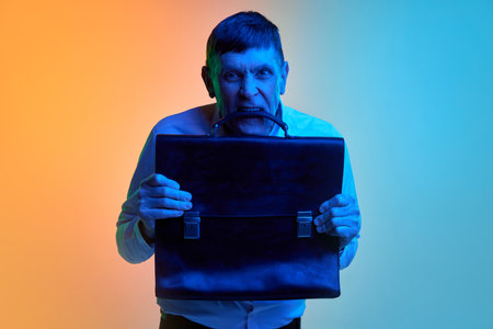 Portrait of mature, middle-aged man emotionally posing with briefcase over gradient blue orange background in neon light. Concept of emotions and lifestyleの写真素材
