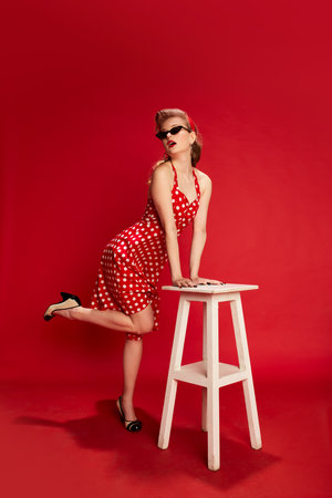 Coquette look. Portrait of beautiful young girl with stylish hairstyle in dress posing against red studio background. Concept of beauty and pin-up styleの写真素材