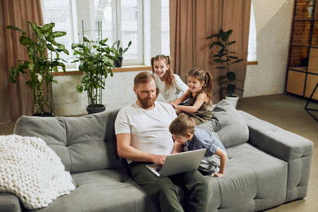 Mature man, father sitting on couch at home and working remotely on laptop with three little kids playing around him and making noiseの写真素材