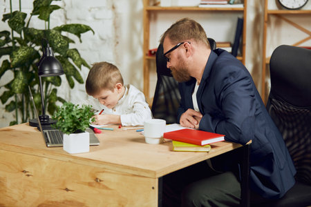 Mature man, father sitting at table, working remotely at home and doing homework with his little sonの写真素材