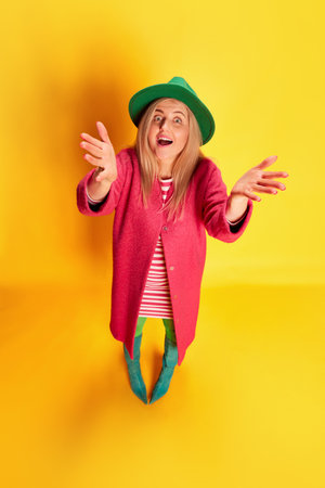 Top view. Portrait of emotive excited woman in colorful clothes, green hat and pink coat posing against yellow studio backgroundの写真素材