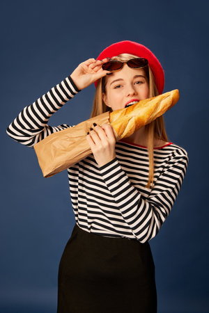 Parisian style. Portrait of beautiful young girl in red beret posing, biting baguette over blue studio background. Concept of fashion, beauty and lifestyleの写真素材