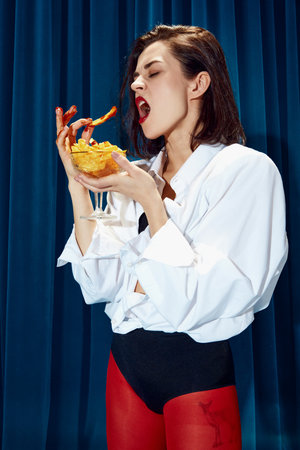 Fast food. Young beautiful brunette woman in white shirt and red tights posing, emotionally eating fries against blue textile backgroundの写真素材