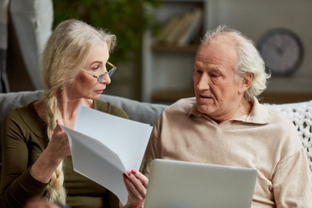 Focused middle aged retired couple, man and woman attentively looking on papers, bills, working on laptop at home. Using internet for paymentの写真素材