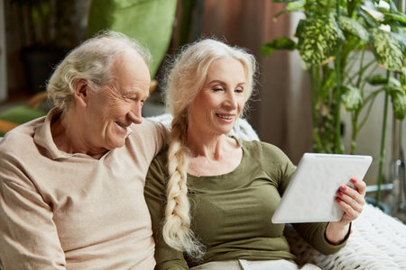Happy smiling people, retired couple, man and woman sitting on couch at home and looking on tablet....