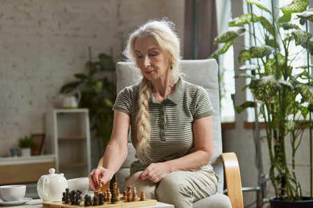 Beautiful senior, retired woman sitting on chair, attentively playing chess. Home interior background. Leisure time and hobbyの写真素材
