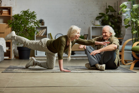 Mature, middle aged woman doing yoga exercises at home with her elderly husband. Couple training together, support and assistanceの写真素材