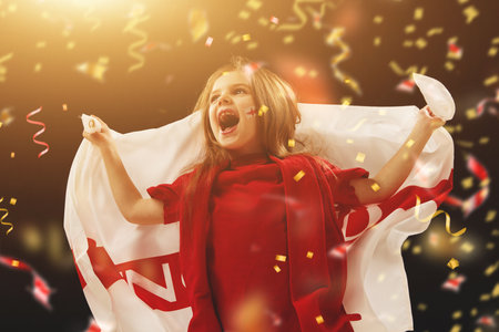 Little girl, happy child with flag of England emotionally watching football match at stadium with confetti. Winning successful gameの写真素材