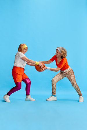 Elderly sportive woman in colorful uniform training, posing, competing for basketball bal against blue studio background. Concept of sport and healthの写真素材
