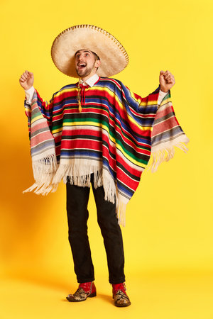 Portrait of young handsome man with happy excited face, in colorful festive clothes, poncho and sombrero posing against yellow studio backgroundの写真素材