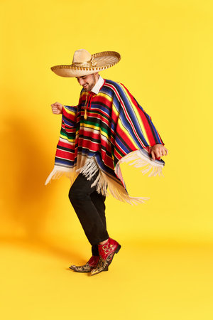 Portrait of young handsome man in colorful festive clothes, poncho and sombrero posing, dancing against yellow studio backgroundの写真素材