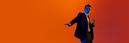 Portrait of young stylish man in jacket and glasses posing, dancing against orange studio background in neon light.の写真素材