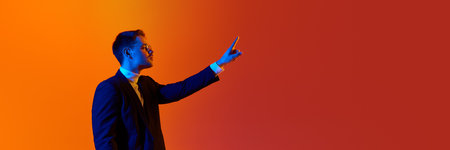 Young man in stylish official jacket and glasses pointing with finger on ilusional screen against orange studio background in neon light.の写真素材