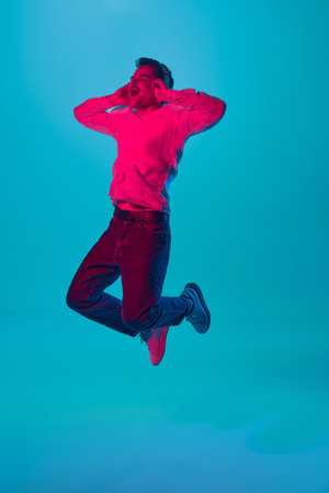 Full-length portrait of young man in white shirt listening to music in headphones and jumping against blue studio background in pink neon light.の写真素材