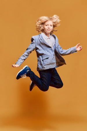 Full-length portrait of little boy, child in jacket jumping, posing against orange studio background. Model with blonde curly hair. Childhood and emotionsの写真素材