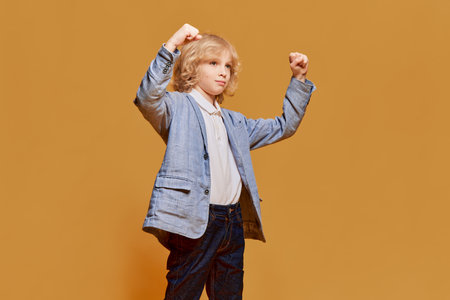Winner. Success. Portrait of little boy, child with blonde curly hair posing in formal wear against orange studio background. Childhood and emotionsの写真素材