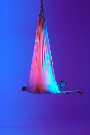 Young man, professional aerial gymnast training with hammock, hanging against gradient blue purple studio background in neon light.の写真素材