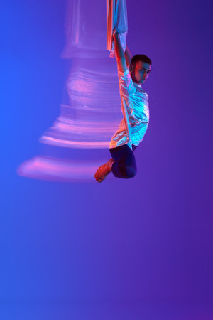 Young man, aerial gymnast, acrobat training with aerial tissue against gradient blue purple background in neon with mixed lightsの写真素材